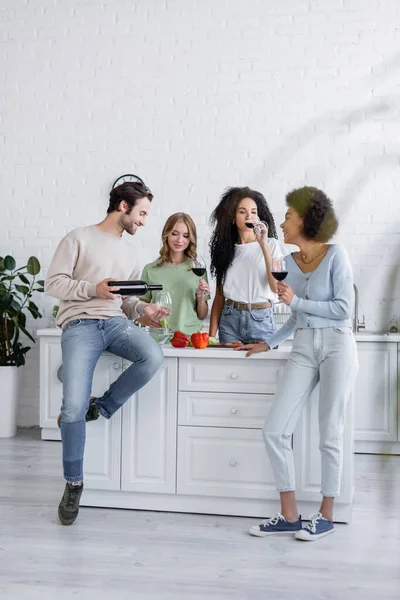 Volle Länge des fröhlichen Mannes, der Rotwein in Glas in der Nähe von jungen interrassischen Frauen in der Küche einschenkt — Stockfoto