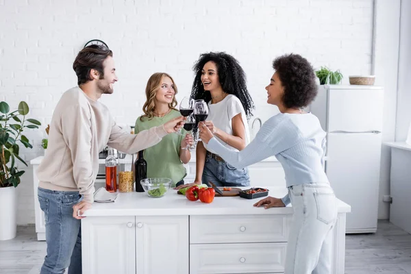 Fröhliche interrassische Freunde klimpern in der Küche mit Rotweingläsern — Stockfoto