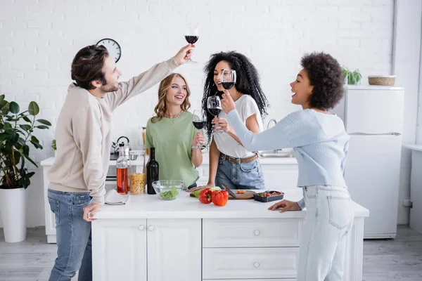 Happy interracial friends holding glasses of red wine in kitchen — Stock Photo