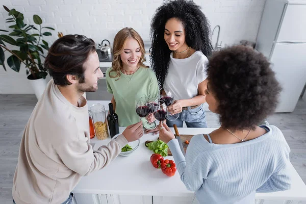 Blick aus der Vogelperspektive auf fröhliche multiethnische Freunde, die in der Küche mit Rotweingläsern klirren — Stockfoto