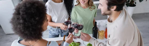 Alegres amigos multiétnicos tintineo vasos de vino tinto en la cocina, pancarta - foto de stock