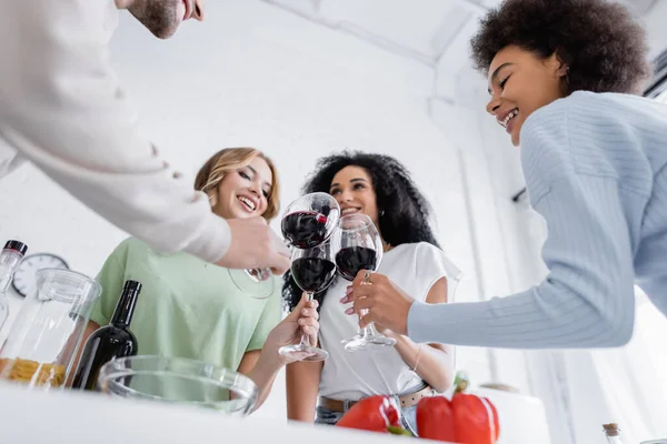 Low angle view of smiling multiethnic friends clinking glasses of red wine in kitchen — Stock Photo