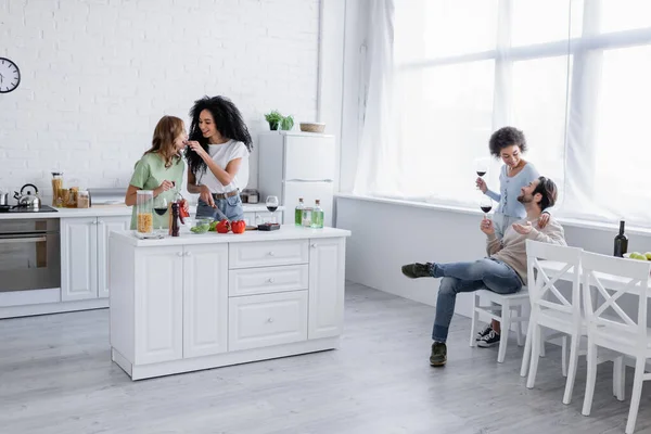 Happy lesbian woman feeding girlfriend near cheerful interracial couple holding glasses on wine — Stock Photo