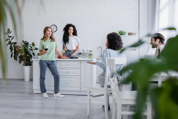 Amis multiethniques joyeux parlant dans la cuisine moderne avec des plantes floues au premier plan — Photo de stock