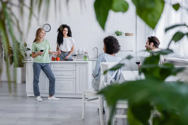 Amigos multiétnicos felices hablando en cocina moderna con plantas borrosas en primer plano - foto de stock