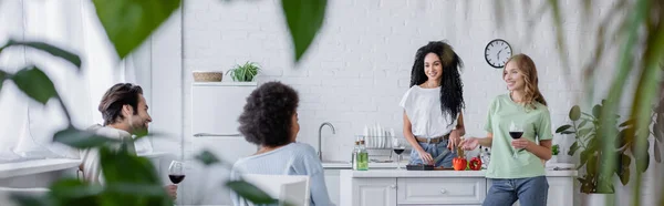 Heureux amis multiethniques parler dans la cuisine moderne avec des plantes floues au premier plan, bannière — Photo de stock