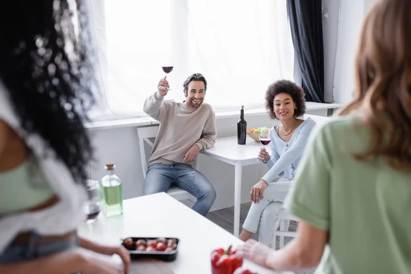 Alegres amigos multiétnicos sosteniendo copas de vino tinto cerca de mujeres borrosas en la cocina moderna - foto de stock