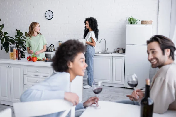 Zufriedenes lesbisches Paar, das sich in der Nähe verschwommener interrassischer Freunde mit Weingläsern anschaut — Stockfoto