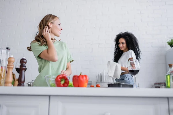 Blonde woman looking at pleased african american girlfriend in kitchen — Stock Photo