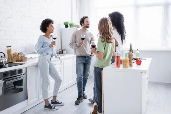 Alegres amigos interracial sosteniendo copas de vino y hablando en la cocina moderna - foto de stock