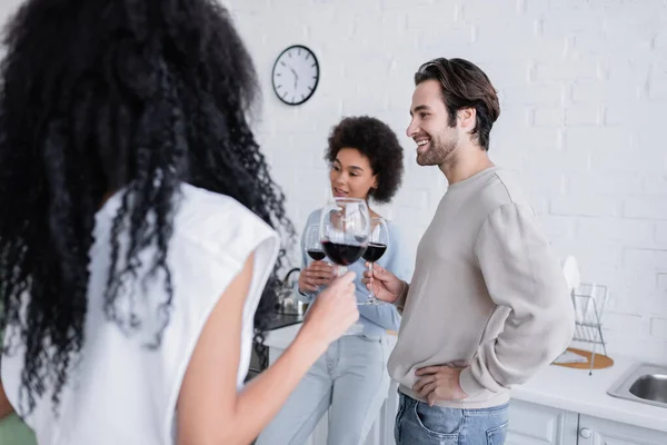 Happy man holding glass of red wine near african american women in kitchen — Stock Photo