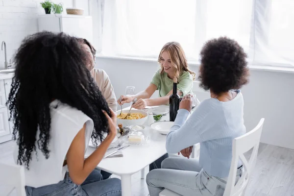 Happy blonde woman serving pasta near interracial friends — Stock Photo