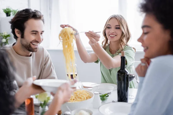 Happy blonde woman serving pasta near man and blurred african american friend — Stock Photo