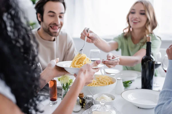 Mujer rubia feliz sirviendo pasta a amigo borroso cerca de hombre alegre - foto de stock