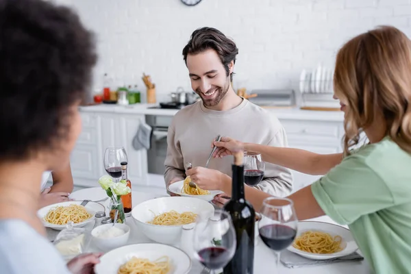 Blonde woman serving pasta to cheerful man near blurred african american friend — Stock Photo