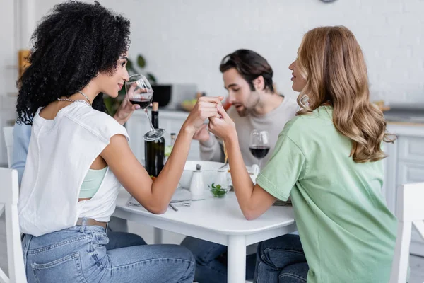 Happy interracial lesbian couple looking at each other and holding hands during lunch with friends — Stock Photo