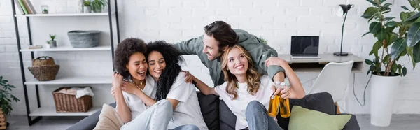 Homme heureux tenant des bouteilles de bière et étreignant les femmes interracial sur le canapé, bannière — Photo de stock