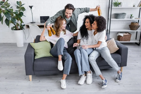 Glücklicher Mann hält Bierflaschen in der Hand und umarmt interrassische Freunde auf dem Sofa — Stockfoto