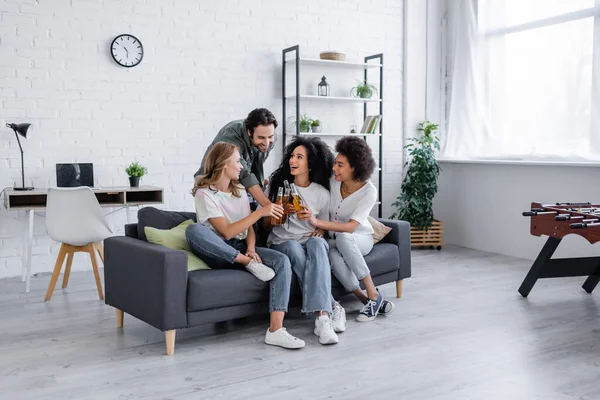 Glücklicher Mann und fröhliche interrassische Frauen, die im Wohnzimmer mit Bierflaschen klingeln — Stockfoto