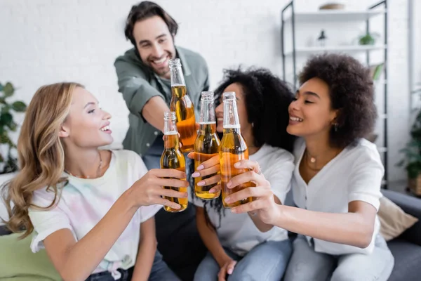 Blurred happy man and joyful interracial women toasting bottles of beer — Stock Photo