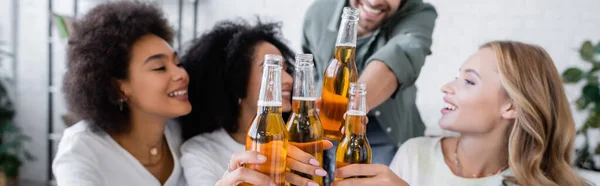 Blurred happy man and joyful interracial women toasting bottles of beer, banner — Stock Photo
