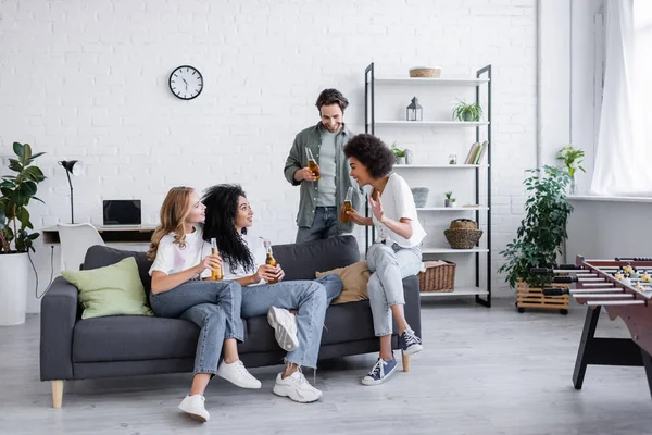 Happy lesbian couple sitting on couch near interracial friends with beer — Stock Photo