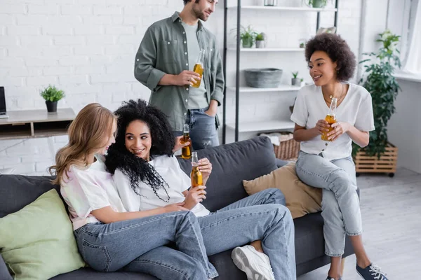 Щаслива лесбіянка пара сидить на дивані біля веселих міжрасових друзів з пляшками пива — стокове фото