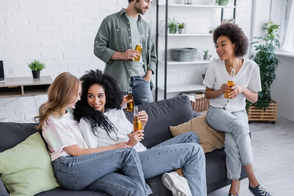 Heureux couple lesbien assis sur canapé près joyeux interracial amis avec bouteilles de bière — Photo de stock