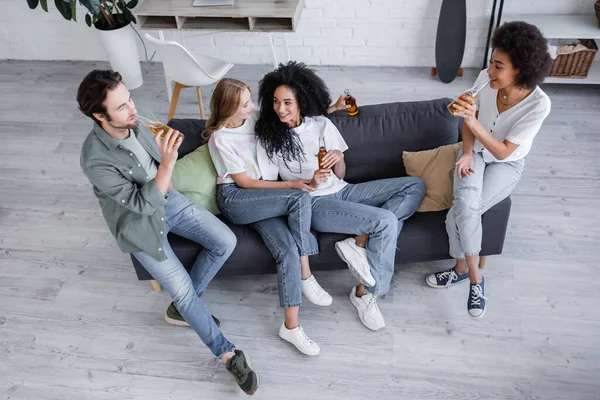 Vista de ángulo alto de feliz pareja lesbiana sentada en el sofá con amigos interracial con cerveza - foto de stock