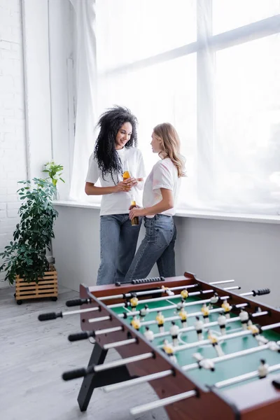 Pareja lesbiana multiétnica sosteniendo botellas de cerveza y de pie cerca de futbolín en la sala de estar - foto de stock