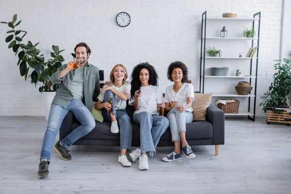 Feliz africano americano mujer clic canales cerca amigos sentado en sofá - foto de stock