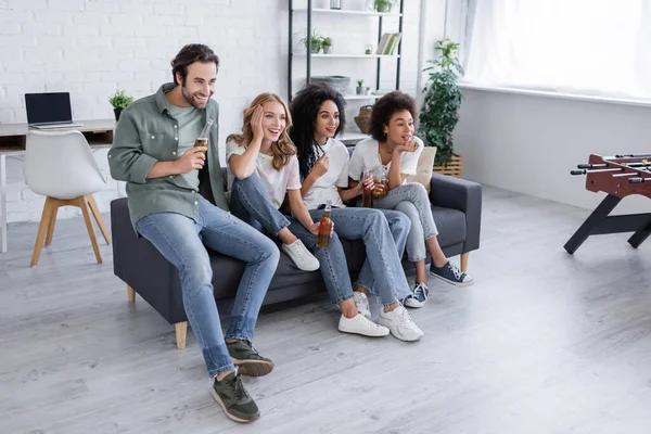 Felici amici interrazziali seduti sul divano, con in mano bottiglie di birra e guardando film — Foto stock