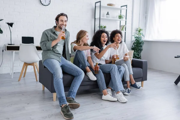 Amis interraciaux joyeux assis sur le canapé, tenant des bouteilles de bière et regardant le film comique — Photo de stock