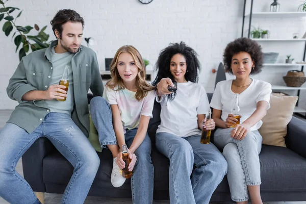 Cheerful african american woman clicking channels near friends sitting on couch — Stock Photo