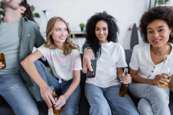 Mujer afroamericana borrosa haciendo clic en canales cerca de amigos felices sentados en el sofá - foto de stock