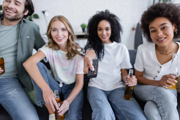 Mujer afroamericana borrosa haciendo clic en canales cerca de amigos alegres sentados en el sofá - foto de stock