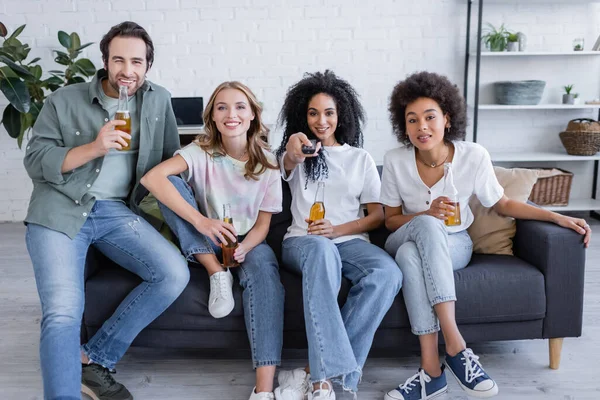 Alegre afro-americana clicando canais perto de amigos felizes sentados no sofá e segurando cerveja na sala de estar — Fotografia de Stock