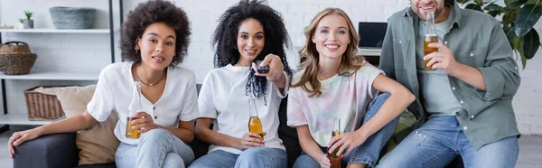 Gioiosa donna afroamericana cliccando canali vicino amici felici seduti sul divano e in possesso di birra, banner — Foto stock