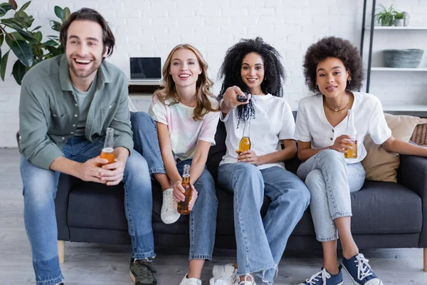 Alegre africano americano mujer clic canales cerca feliz amigos sentado en sofá y celebración de cerveza en sala de estar - foto de stock