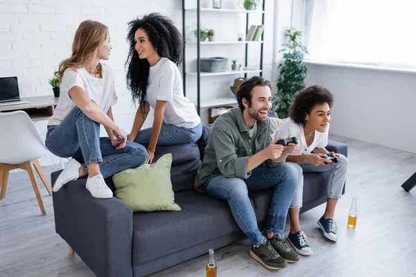 KIEW, UKRAINE - 7. OKTOBER 2021: glückliches multiethnisches lesbisches Paar sitzt auf Couch in der Nähe von Freunden und spielt Video — Stockfoto