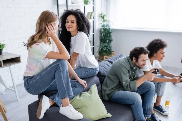 QUIIV, UCRÂNIA - OUTUBRO 7, 2021: alegre casal lésbico multiétnico sentado no sofá perto de amigos tensos jogando vídeo — Fotografia de Stock