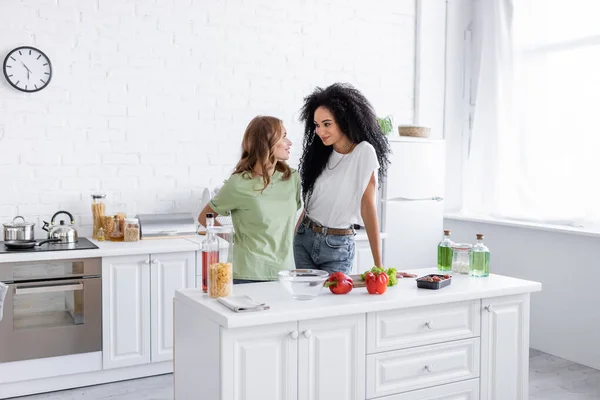 Feliz pareja de lesbianas multiétnicas mirándose en la cocina moderna - foto de stock