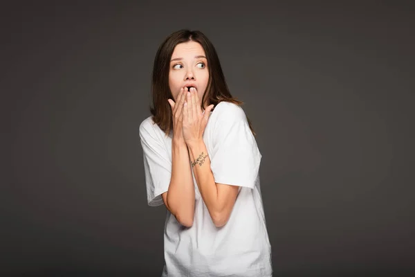Scared and shocked woman covering mouth with hands while looking away isolated on dark grey — Stock Photo