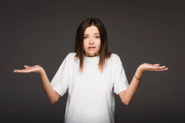 Femme embarrassée regardant la caméra et montrant un geste d'haussement d'épaules isolé sur gris foncé — Photo de stock