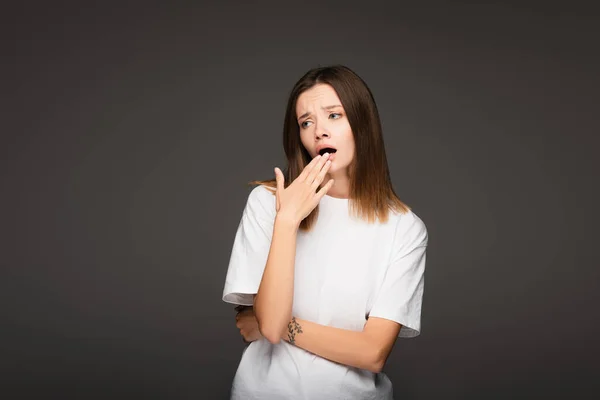 Bored woman in white t-shirt covering mouth with hand while yawning isolated on dark grey — Stock Photo