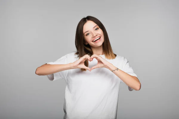 Felice giovane donna che mostra il segno del cuore mentre guarda la fotocamera isolata sul grigio — Foto stock