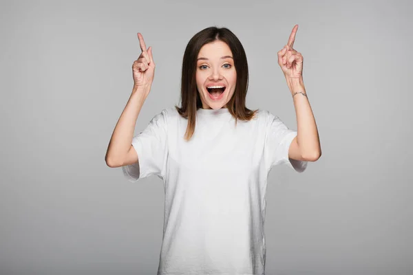Amazed woman looking at camera and pointing up with fingers isolated on grey — Stock Photo
