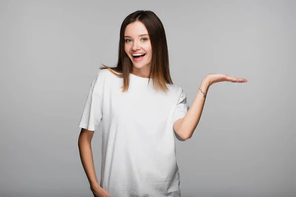 Femme heureuse en t-shirt blanc regardant caméra et pointant avec la main isolé sur gris — Photo de stock