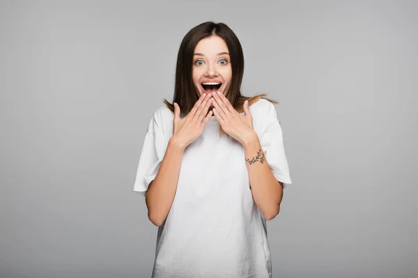 Amazed and happy woman holding hands near open mouth isolated on grey — Stock Photo