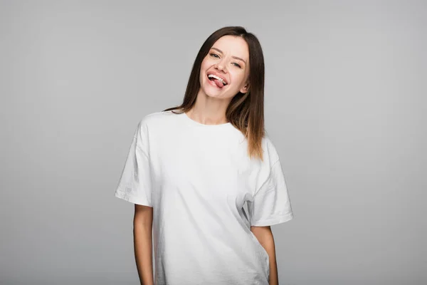 Cheerful woman in white t-shirt sticking out tongue isolated on grey — Stock Photo